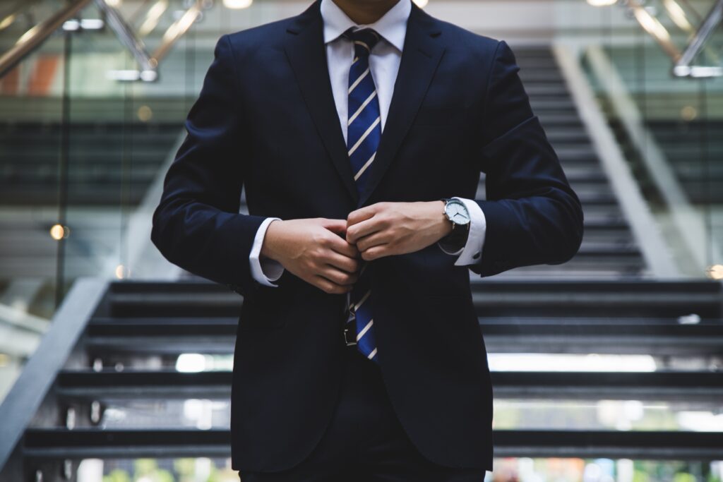 A business man in a dark blue suit with a white and bright blue striped tie buttoning his shirt as he walks down a flight of stairs